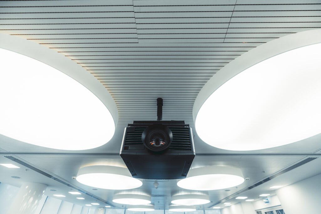 Wide-angle view of a black modern projector with a plastic case and a huge lens with aberrations on it, mounted to the ceiling with multiple large round ceiling lamps lighting a modern conference room